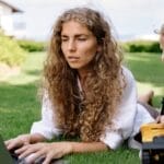 Woman working on a laptop while lying on grass with her child nearby playing.