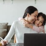 Happy young Asian woman working remotely from home with laptop and tablet while adorable little daughter hugging from behind