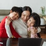 A mother embraces her children while working from home, blending family time with remote work.