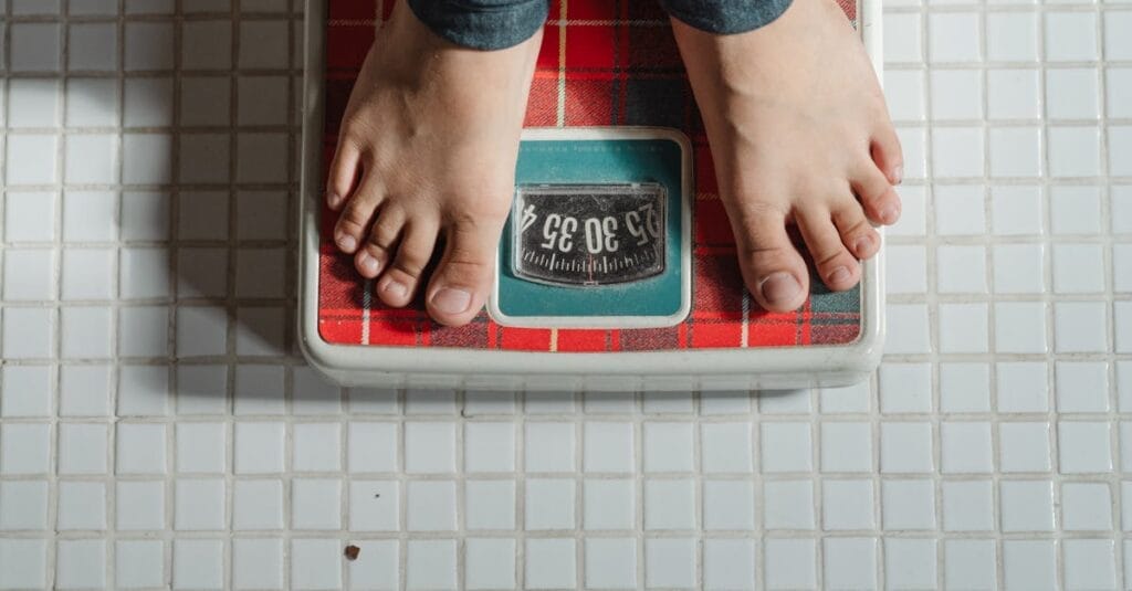 From above crop anonymous barefoot child in jeans standing on weigh scales on tiled floor of bathroom