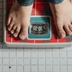 From above crop anonymous barefoot child in jeans standing on weigh scales on tiled floor of bathroom