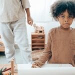 Modest black kid in casual clothes sitting at table with stationery and paper close to faceless sibling and looking at camera