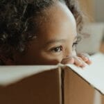 A charming portrait of a child with curly hair peeking out from a cardboard box indoors.