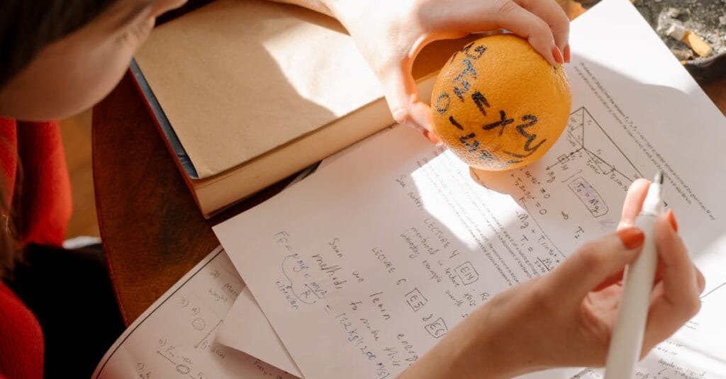 Student studying math creatively using an orange and notes in warm indoor light.