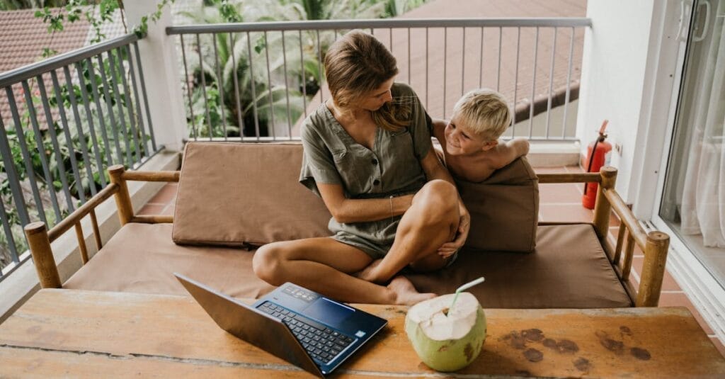 A mother and son relaxing on a balcony with a laptop and coconut nearby.