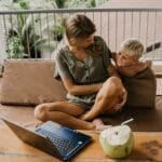 A mother and son relaxing on a balcony with a laptop and coconut nearby.