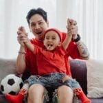 Father and son bonding on the couch, dressed in red, enjoying a football moment together.