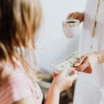 A woman hands a dollar bill to her daughter while holding a coffee cup, indoors.