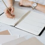 A woman writes in a planner on a marble table, surrounded by swatches. Creative and organized workspace.