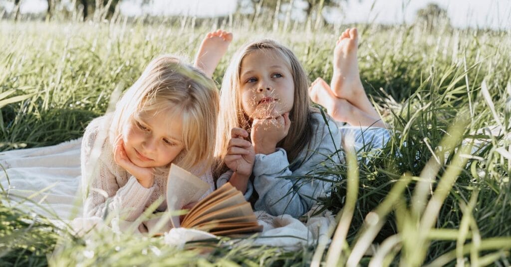 Two young girls lying on a picnic blanket in a sunny field, reading and daydreaming.