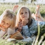 Two young girls lying on a picnic blanket in a sunny field, reading and daydreaming.