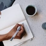 Person writing in a planner with coffee at a café, focusing on planning and productivity.