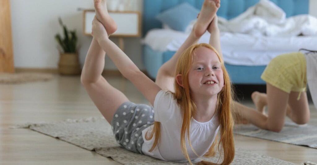 Full body of cheerful girl in casual clothing with reddish hair looking away with smile while stretching legs on carpet in bright cozy living room on blurred background