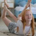 Full body of cheerful girl in casual clothing with reddish hair looking away with smile while stretching legs on carpet in bright cozy living room on blurred background