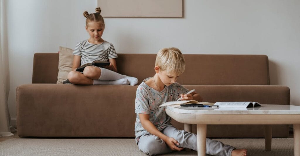 A boy studies while a girl relaxes on the sofa, both in a comfortable indoor room setting.