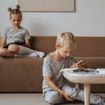 A boy studies while a girl relaxes on the sofa, both in a comfortable indoor room setting.