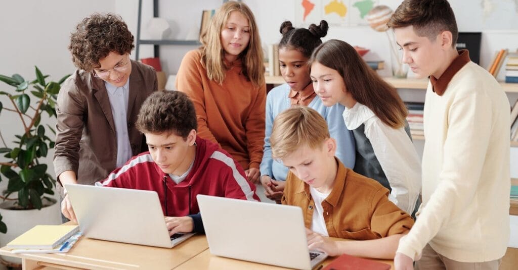 A group of diverse students collaborating on laptops in a classroom setting, guided by a teacher.