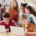 A group of diverse students collaborating on laptops in a classroom setting, guided by a teacher.