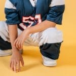 Young child in football gear squatting on a yellow background, showcasing sporty attire and enthusiasm.