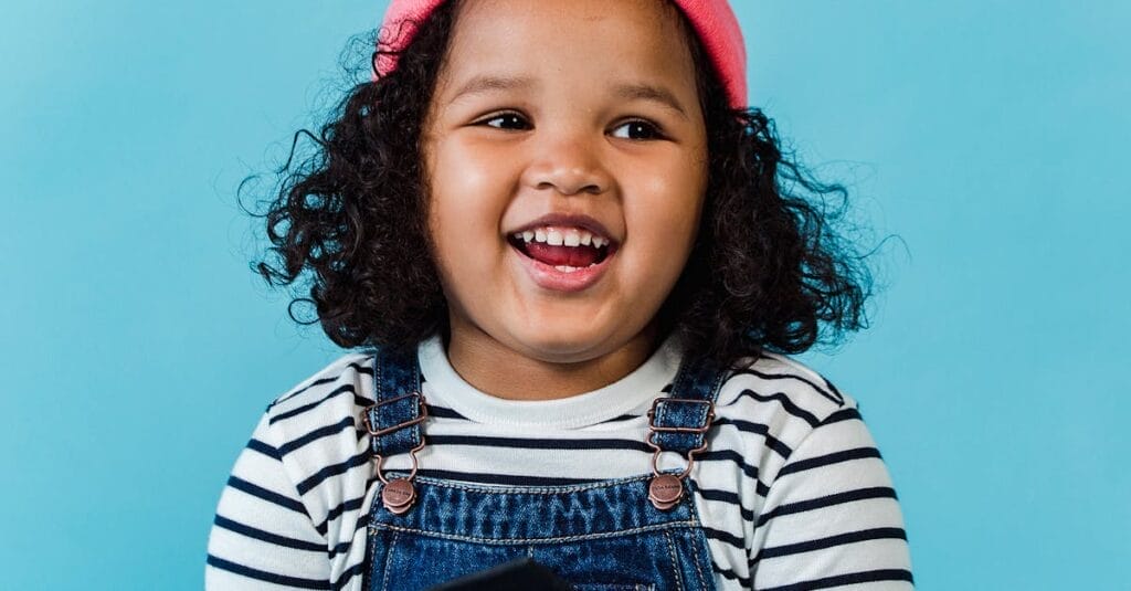 A smiling young girl in a striped shirt and pink beanie enjoying a book against blue background.