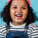 A smiling young girl in a striped shirt and pink beanie enjoying a book against blue background.