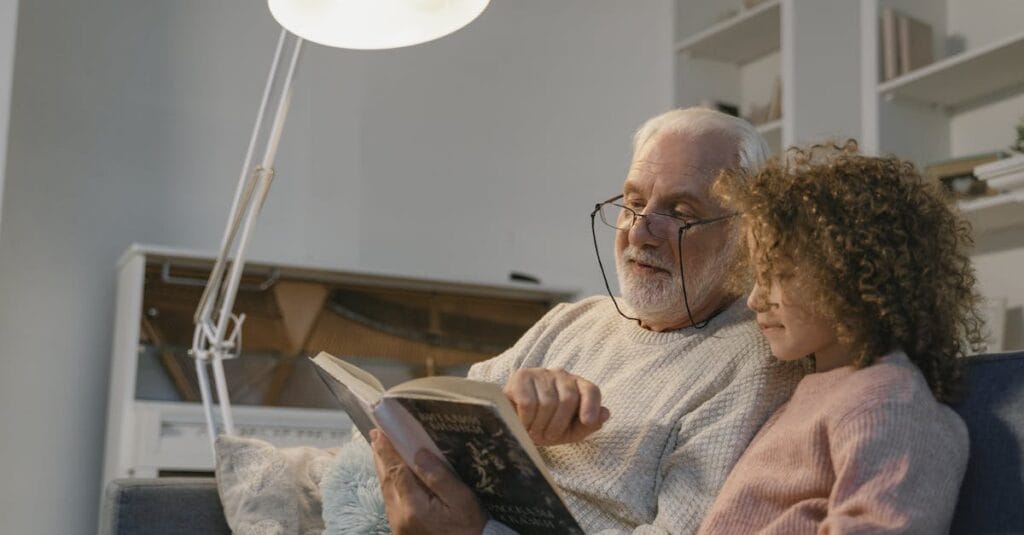 An elderly man reading a book with his granddaughter in a cozy indoor setting.
