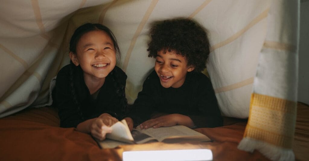 Two children smiling and reading a book in a cozy blanket fort indoors.