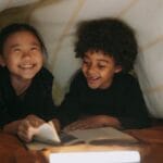 Two children smiling and reading a book in a cozy blanket fort indoors.