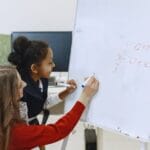 A teacher helps a student with math on a whiteboard in a classroom setting.