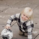 Little blond child in warm jumper playing with ball crawling on ground on play yard and looking at camera