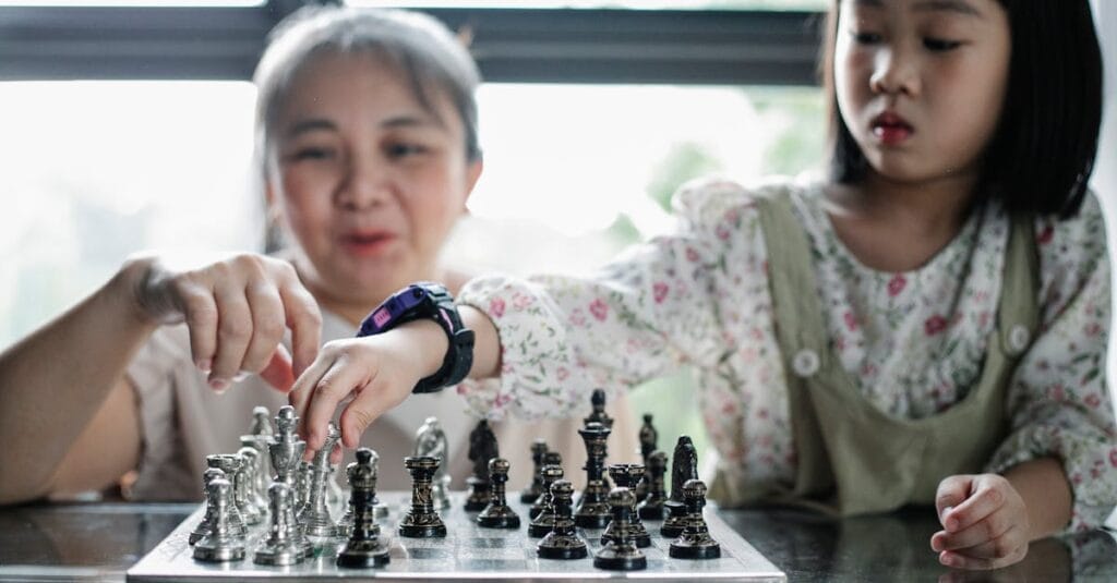 Calm Asian babysitter and girl playing chess at glass table in light room