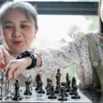 Calm Asian babysitter and girl playing chess at glass table in light room