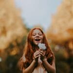 Smiling redheaded girl enjoys nature holding a flower outdoors during the day.