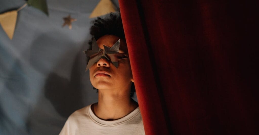 A child wearing paper star glasses peeks behind a red curtain, playing pretend.