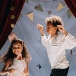 Two joyful kids performing in a homemade theater setup with costumes and curtains, having a fun time indoors.