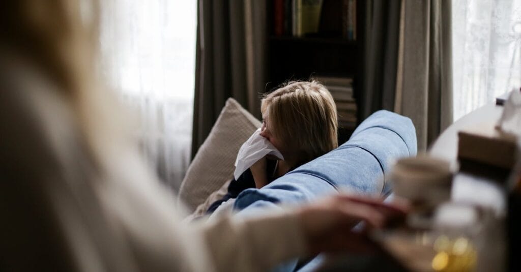 A young child sits on a couch indoors, using a tissue, suggesting illness or allergy.