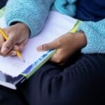 Child sitting outdoors, writing math homework in a notebook with concentration.