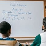 Cheerful African American female math teacher in formal clothes standing near whiteboard and explaining lecture task to multiracial pupils