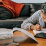 A young Asian girl with pigtails diligently completing homework at home on the sofa.