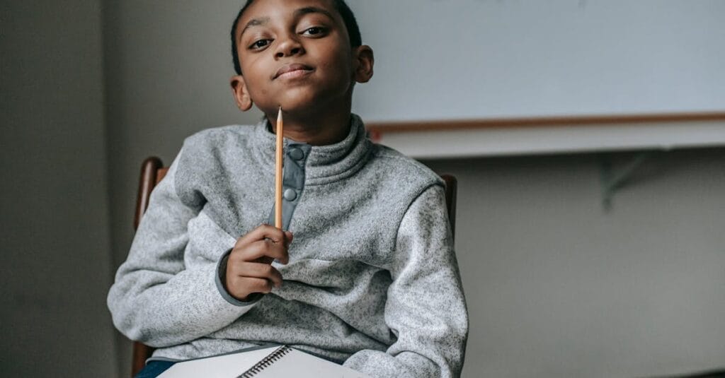 Cute little black boy wearing fleece sweater sitting on chair with pencil under chin and dreamy looking at camera during break after lesson in school