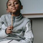 Cute little black boy wearing fleece sweater sitting on chair with pencil under chin and dreamy looking at camera during break after lesson in school