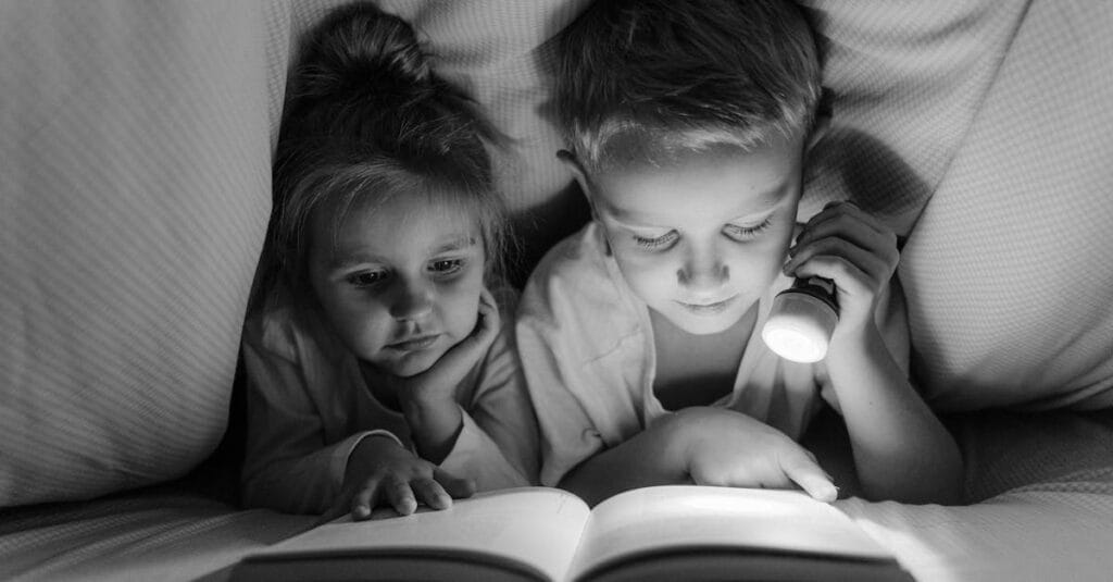 A boy and girl reading with a flashlight under a blanket in grayscale