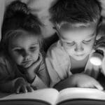 A boy and girl reading with a flashlight under a blanket in grayscale