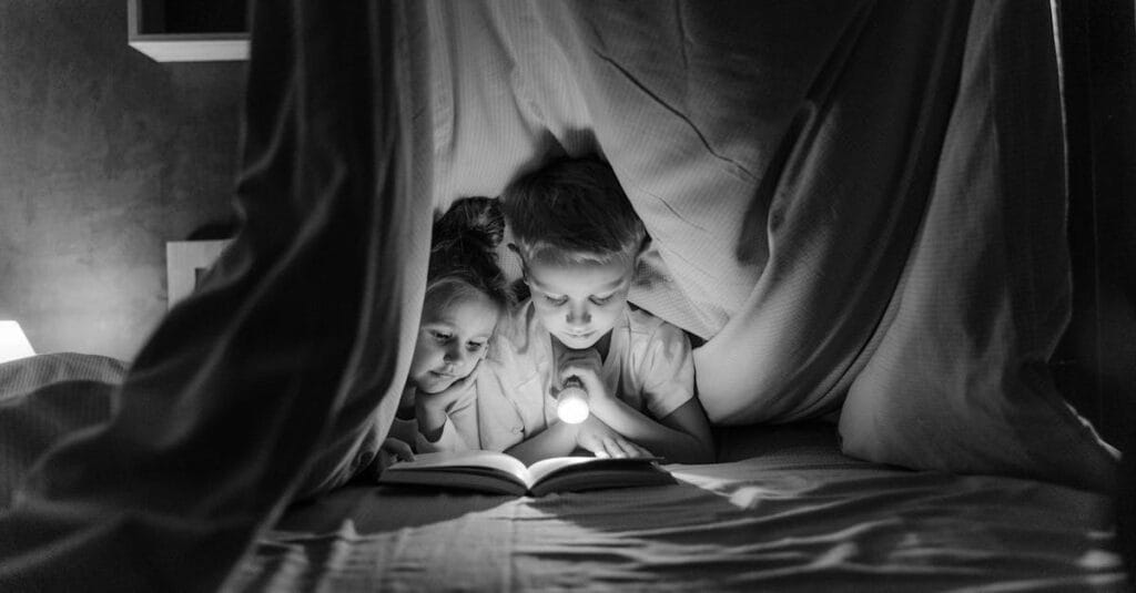 Two children reading a book with a flashlight under a cozy blanket fort at night.