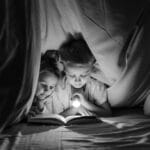 Two children reading a book with a flashlight under a cozy blanket fort at night.