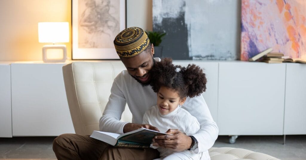 Positive young African American father in hat reading book to cute little daughter sitting on armchair at home