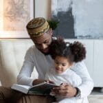 Positive young African American father in hat reading book to cute little daughter sitting on armchair at home