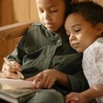 Two siblings enjoy a cozy indoor moment, writing and bonding in a warm setting.