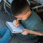 Child sitting on sidewalk, concentrating on writing in notebook with pencil, surrounded by urban graffiti.