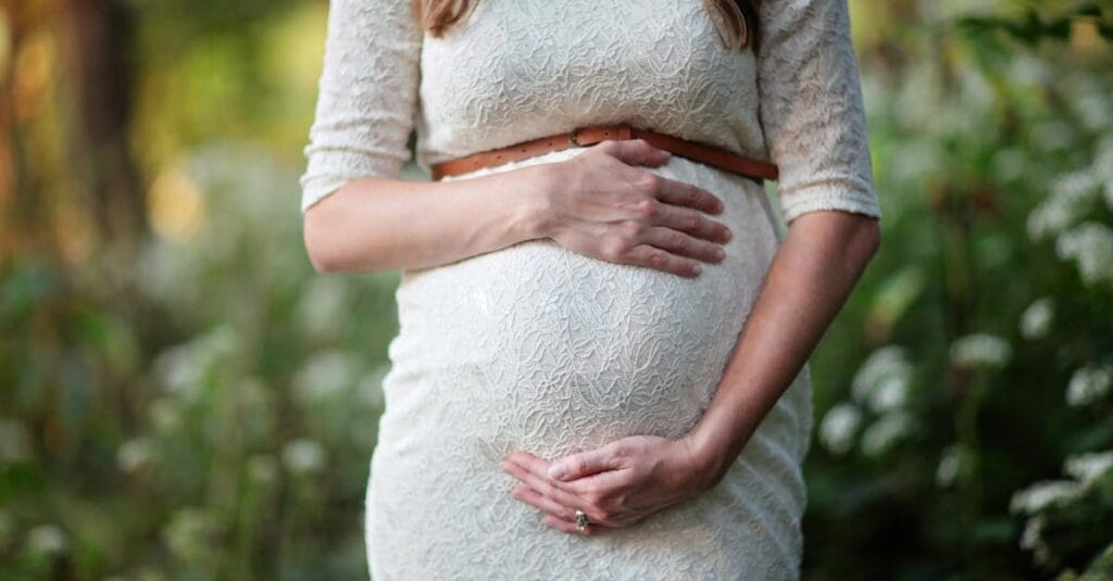 A pregnant woman in a lace dress gently cradling her belly in an outdoor setting.